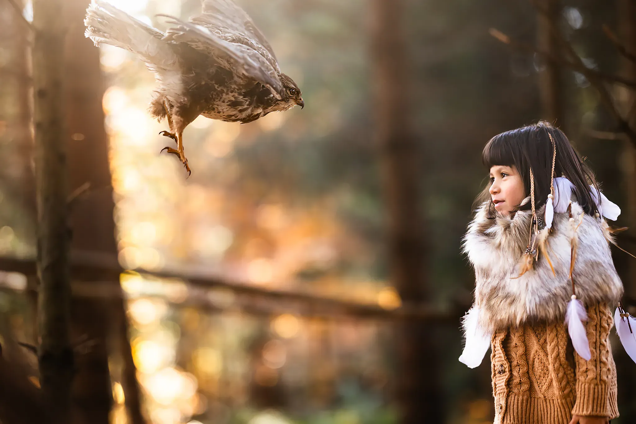 Une enfant en manteau de fourrure et plumes dans les cheveux regarde un rapace voler vers elle, dans une forêt à contre-jour